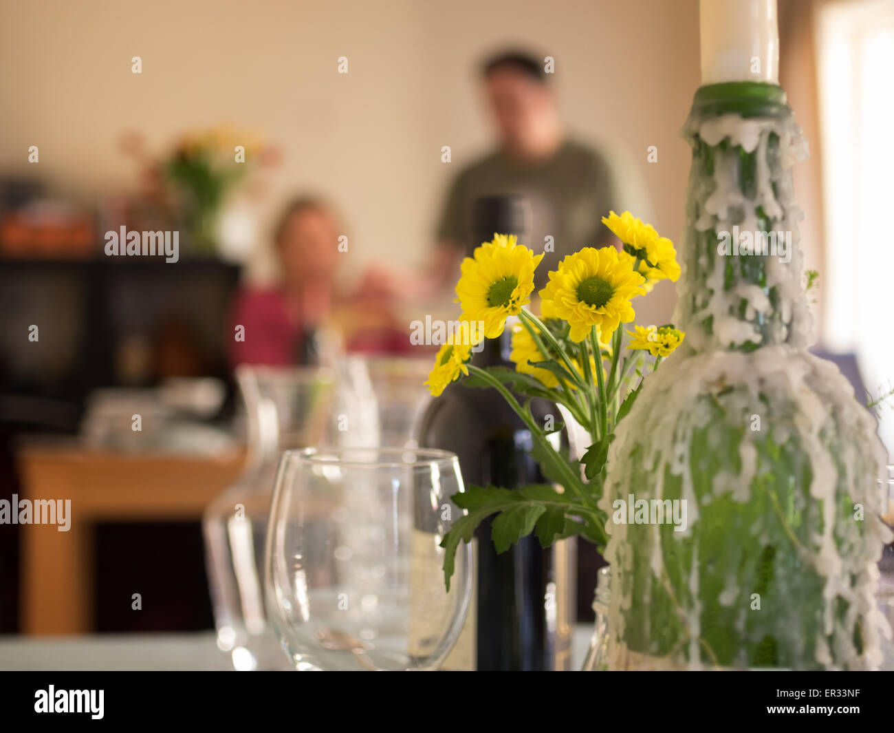 Two people preparing for supper club dinner party. Candle wax encrusted bottles, wine glasses and yellow flowers in foreground. Stock Photo