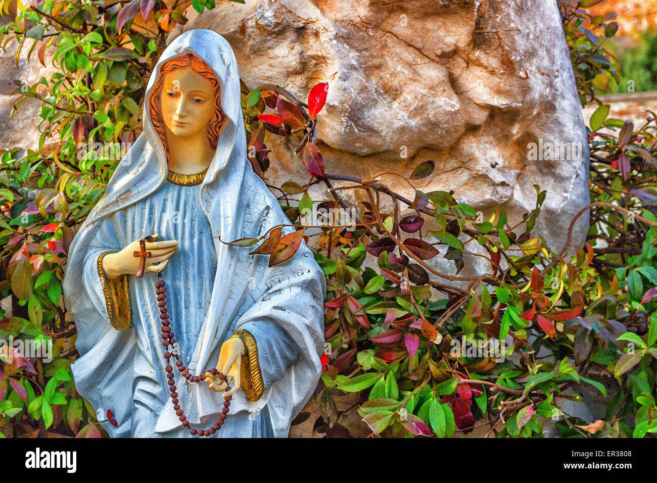 Statue of the Blessed Virgin Mary with wooden prayer beads necklace in a house rock  garden  with treesin  Medjugorje Stock Photo