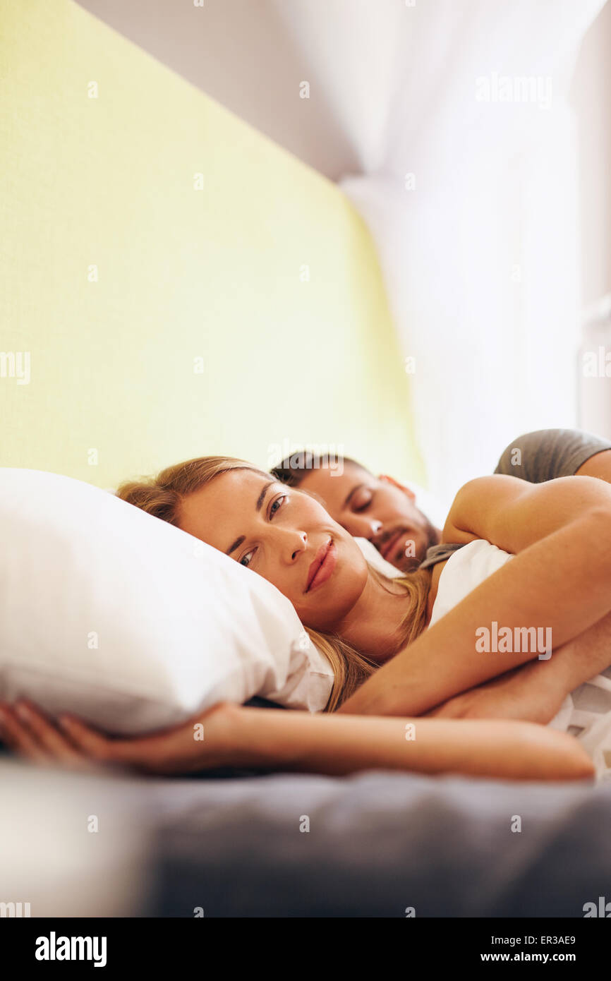 Young couple lying together on bed. Beautiful young woman lying on bed and looking at camera with her husband sleeping. Stock Photo
