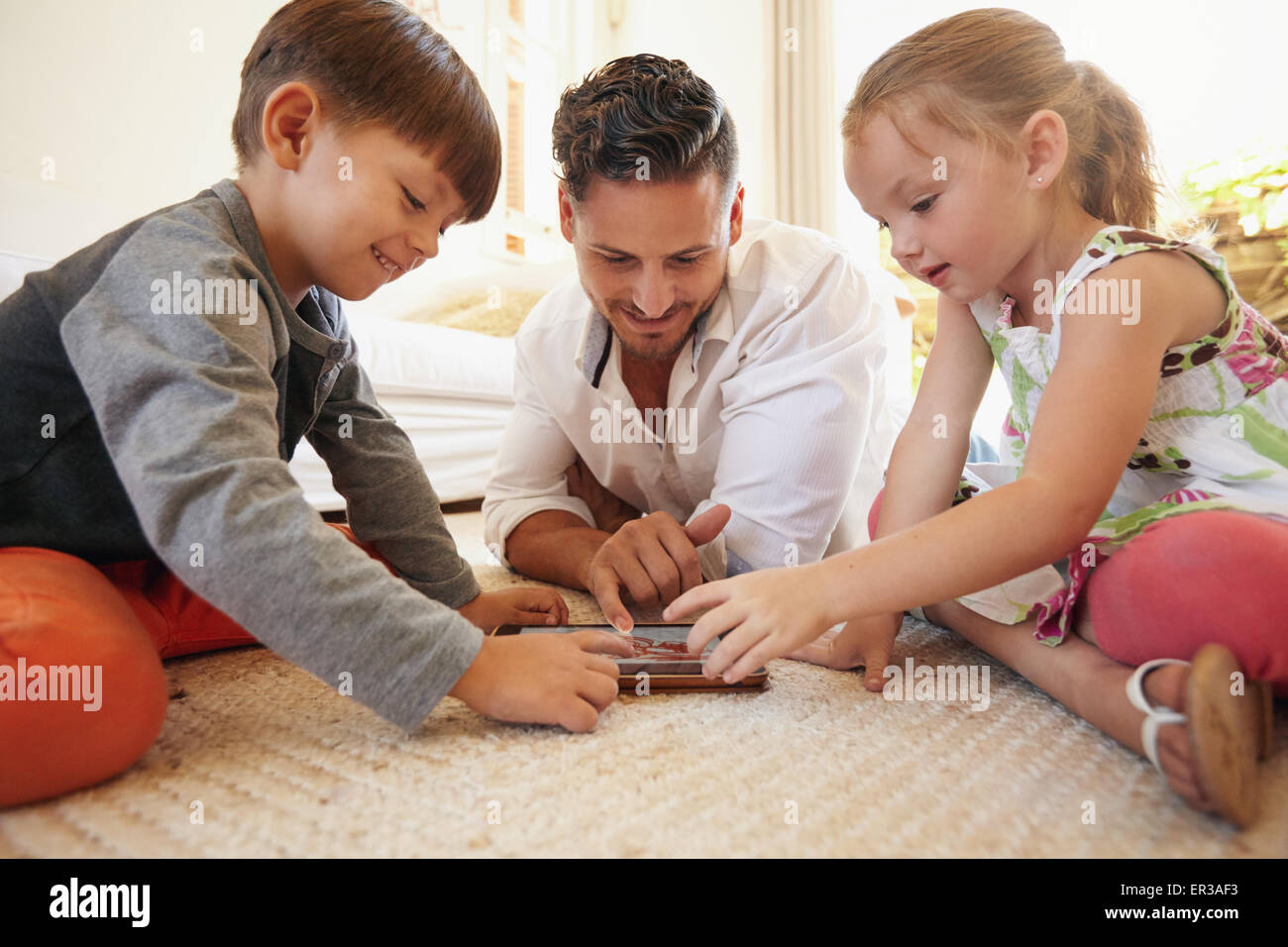 Father with son and daughter sitting on floor using digital tablet indoors. Happy young family together at home using touchpad c Stock Photo