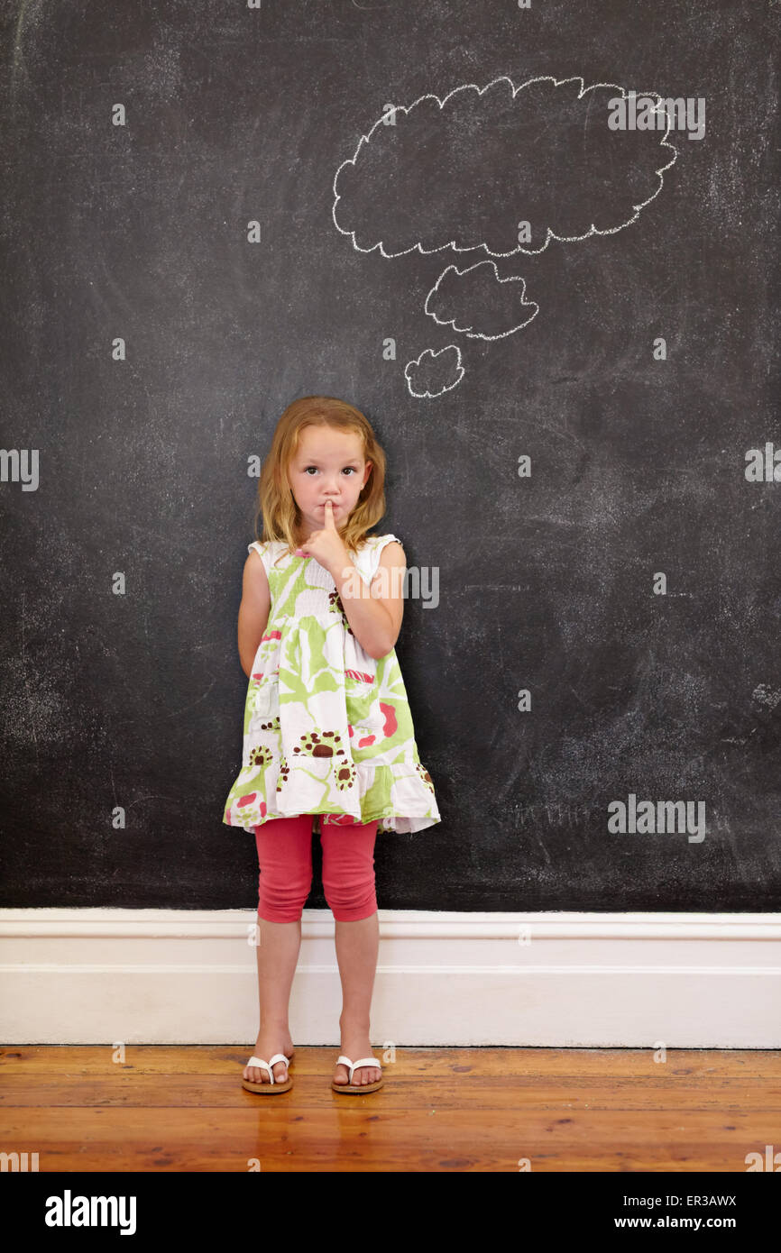 Cute little girl with her finger on lips and a thought bubble on blackboard. Little girl doing a please keep quiet gesture towar Stock Photo