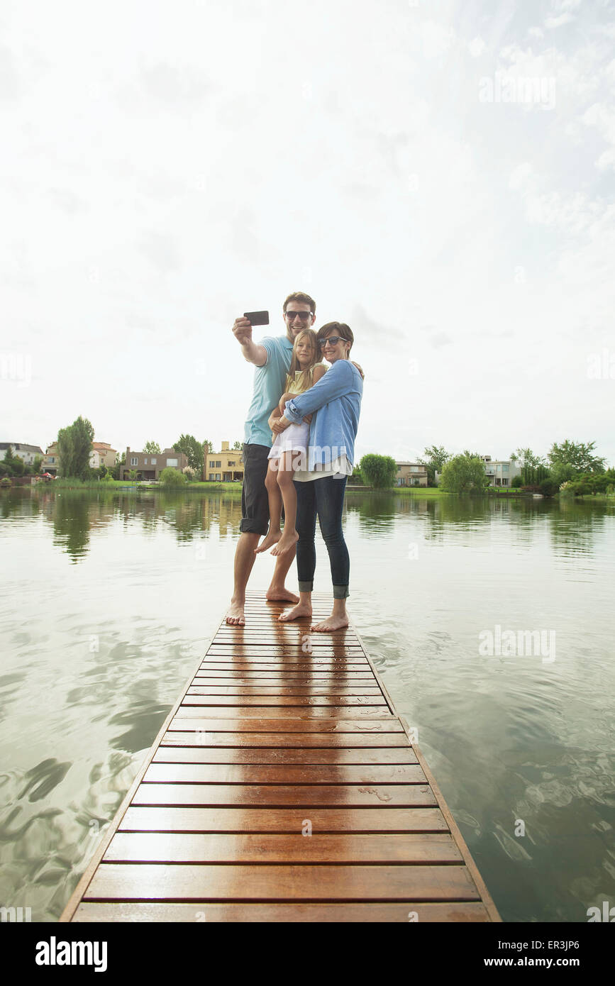 Family with young daughter standing together at end of lake pier taking selfie Stock Photo