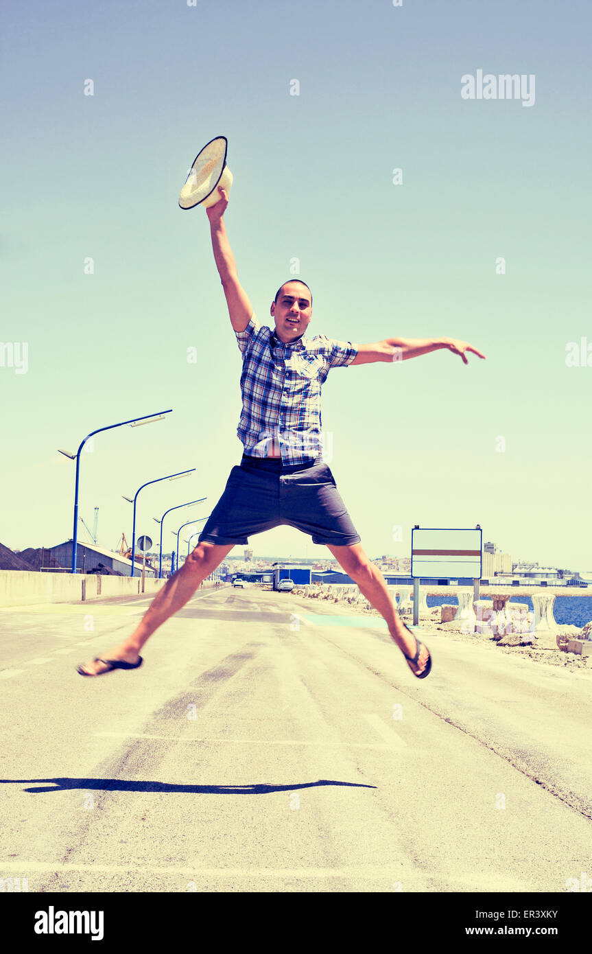a young caucasian man with a straw hat in his hand jumping in a no traffic road near the sea Stock Photo