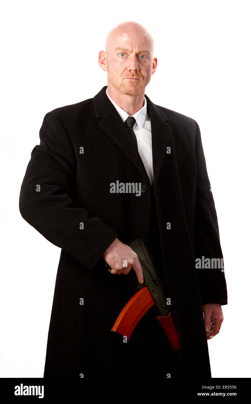 bald headed man wearing heavy black overcoat holding ak-47 against a white studio background model released image Stock Photo