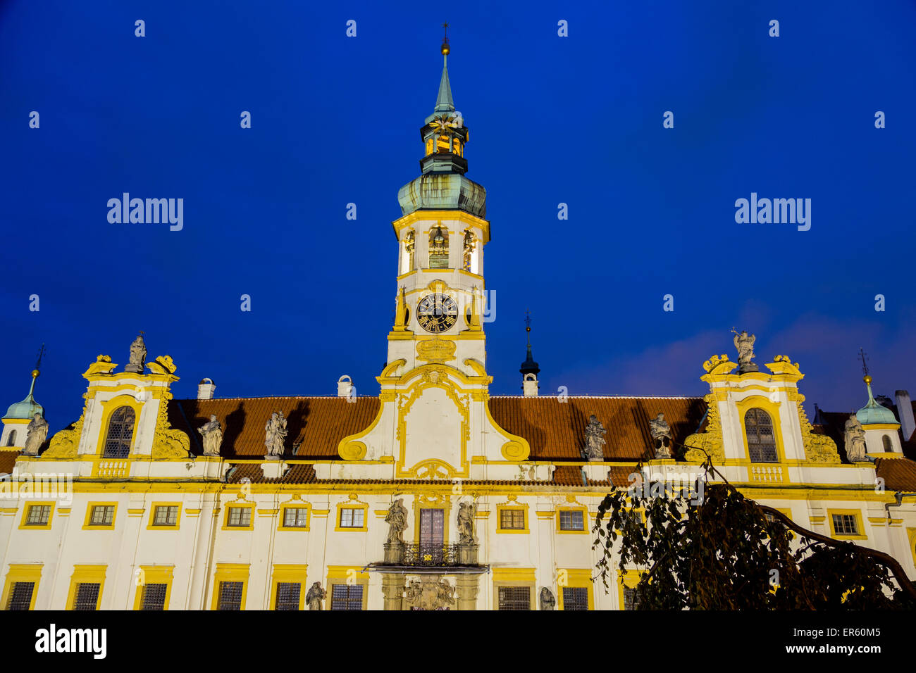 Night view of facade of Loreta church in Prague: green rooftop on white belfry, white walls, red rooftop. Stock Photo