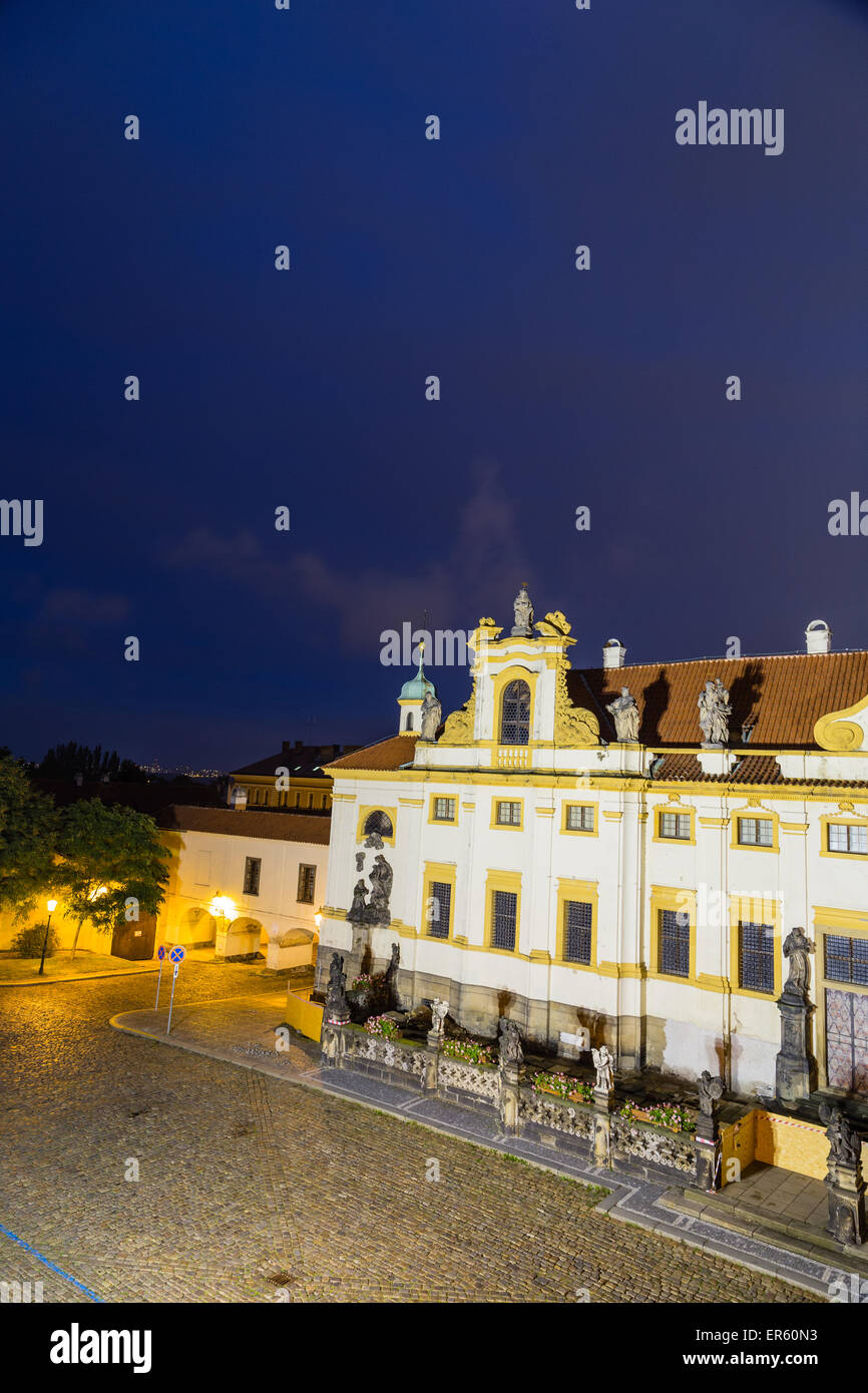 Night view of facade of Loreta church in Prague: green rooftop on white belfry, white walls, red rooftop. Stock Photo