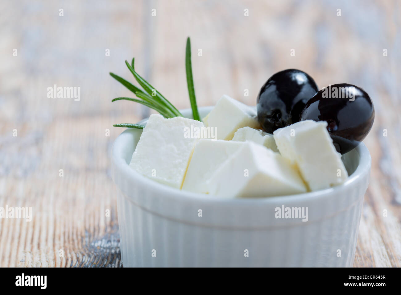 Feta slices with olives and rosemary in a white bowl on a rustic wooden table Stock Photo