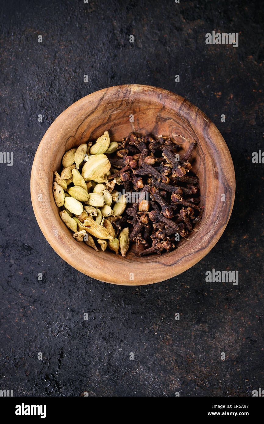 Cardamom seeds and cloves in olive wood bowl over black background. Top