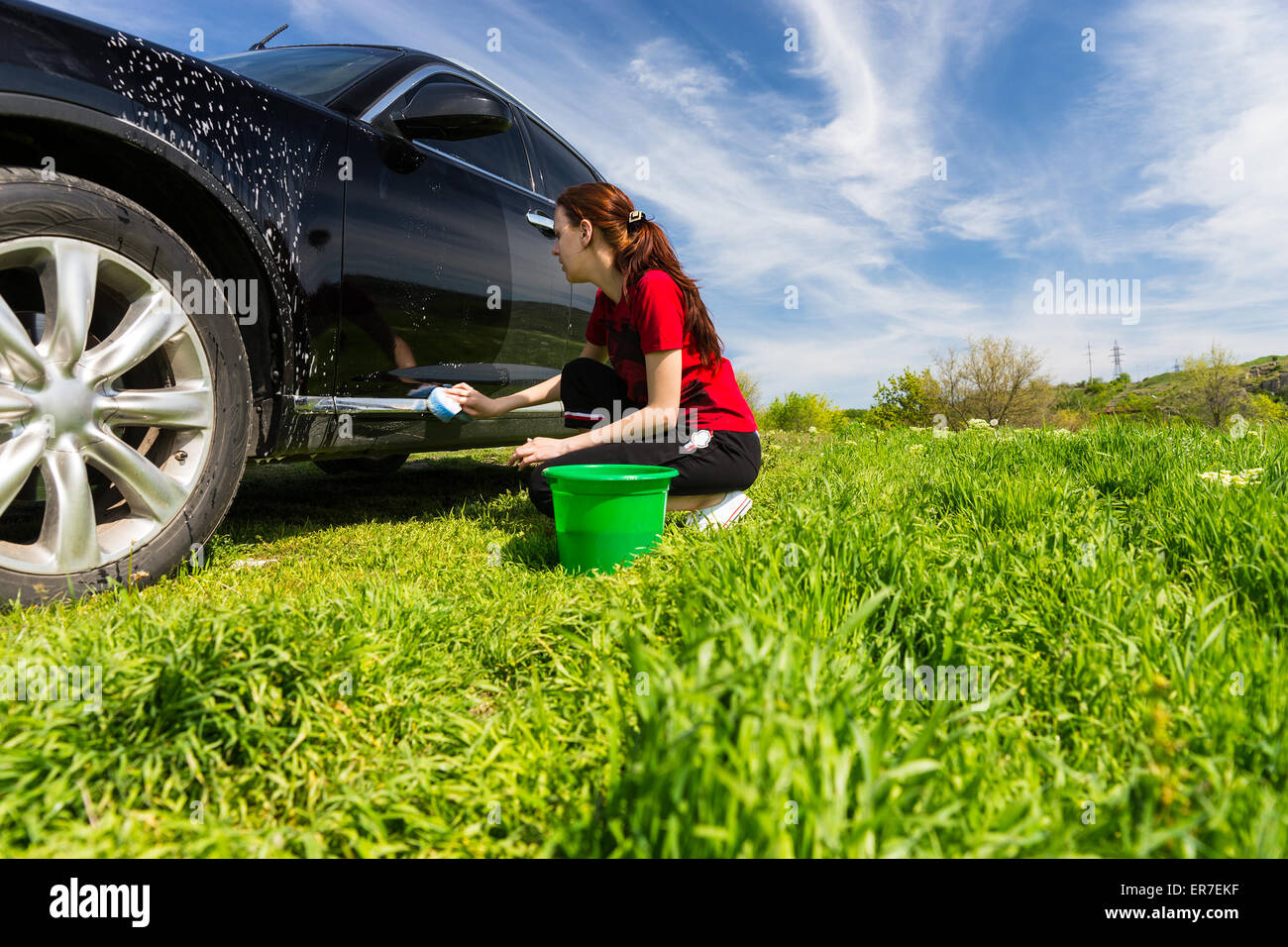 Woman Wearing Red T-Shirt Washing Black Vehicle in Field, Crouching Next to Green Bucket Wringing Soapy Sponge on Bright Sunny Day. Stock Photo
