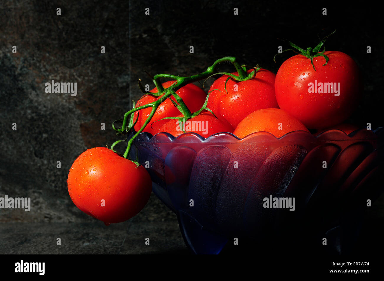 Still life closeup of bright red tomatoes in blue vintage bowl against a dramatic black slate kitchen setting Stock Photo