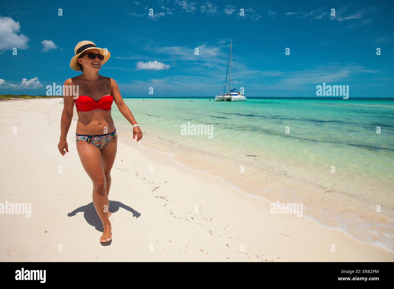 A young woman wearing a bikini and sun hat walks a sandy beach. Stock Photo