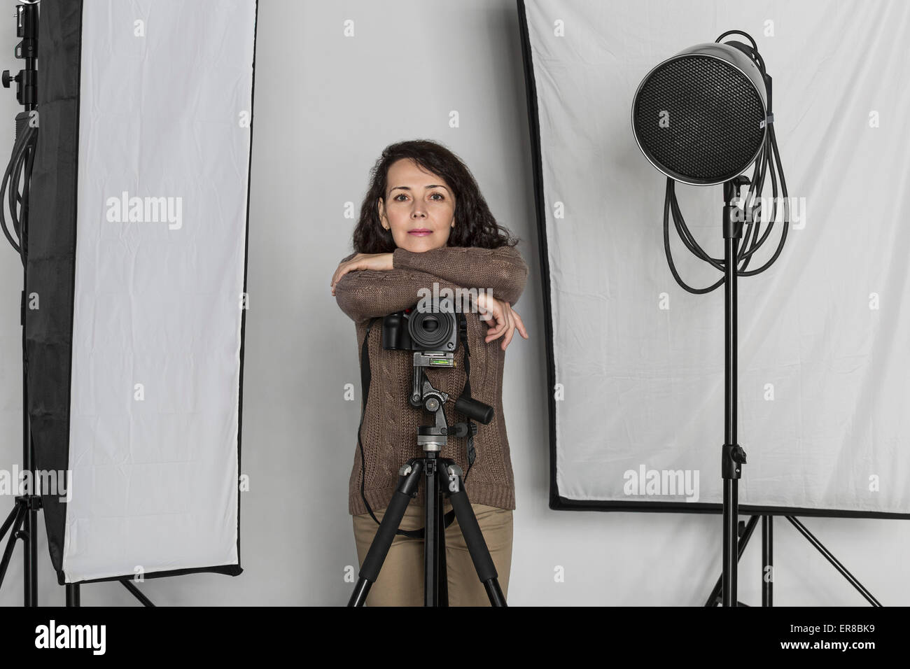 Portrait of confident mature female photographer leaning on camera tripod in photo studio Stock Photo