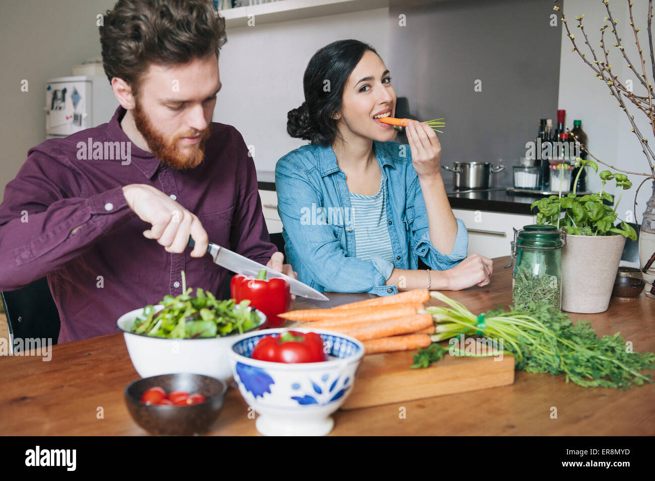 Young man chopping red bell pepper beside woman eating carrot at kitchen table Stock Photo