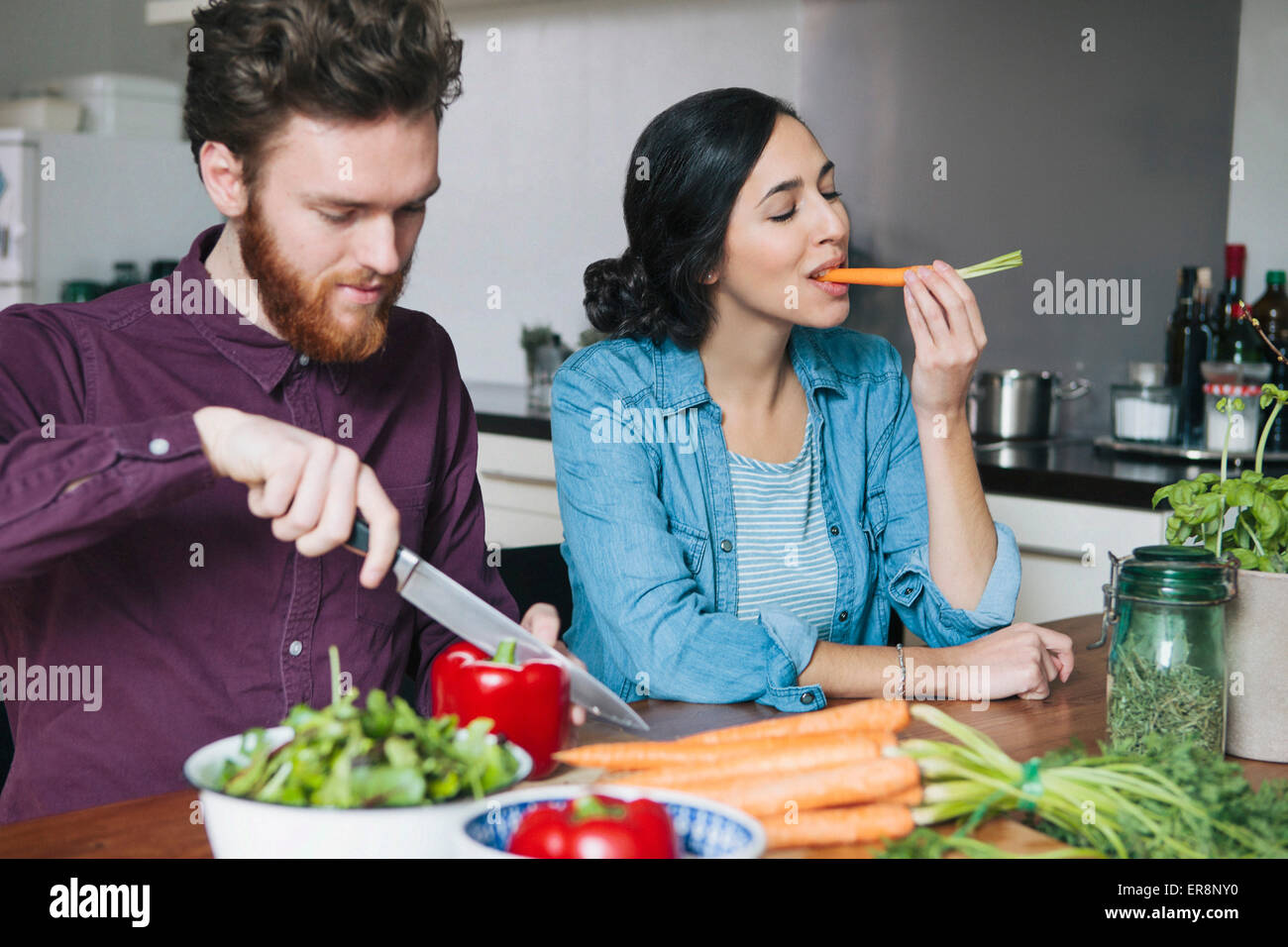 Young man chopping red bell pepper beside woman eating carrot at kitchen table Stock Photo