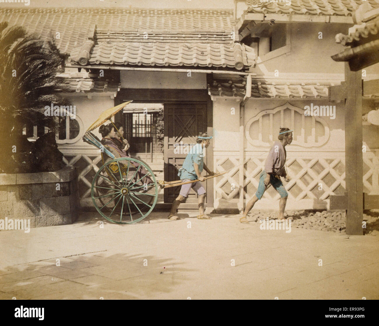 Two women riding in a rickshaw. Photograph shows two women with a large parasol riding in a rickshaw pulled by a man with a second man walking nearby, possibly to relieve the first man. Exterior view of building with tiled roof in the background. Date ca. Stock Photo