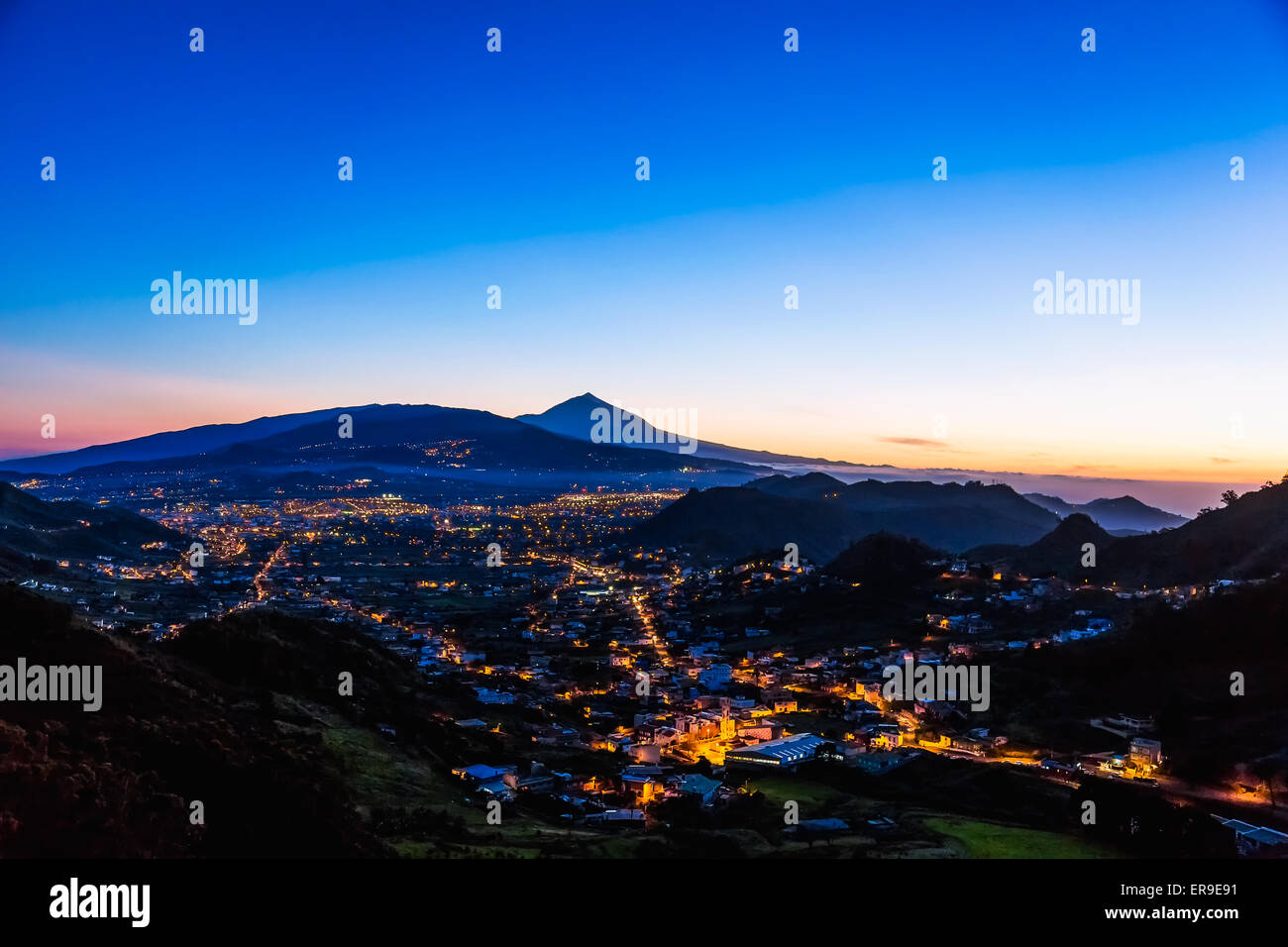 City or town with illumination after sunset or sundown at evening in mountains with blue sky and Teide volcano on background Stock Photo