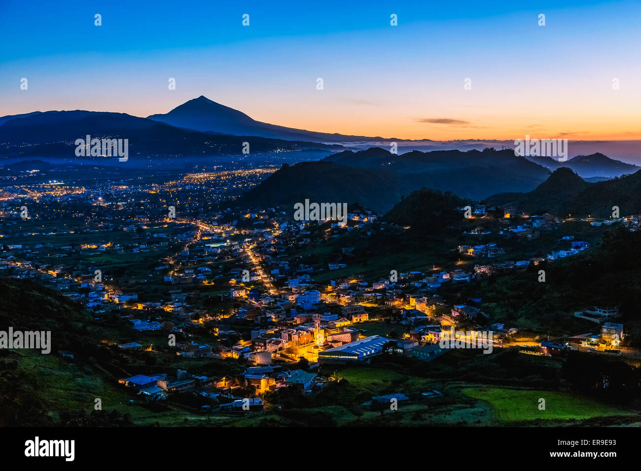 City or town with illumination after sunset or sundown at evening in mountains with blue sky and Teide volcano on background Stock Photo