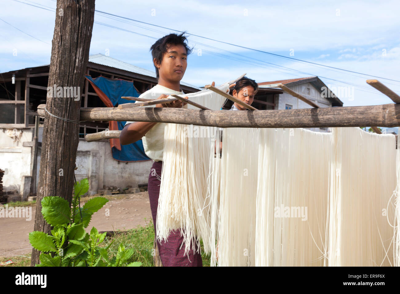 In Hsipaw, Burma, fresh-made rice noodles are hung up on poles to dry in the open air after being made in the buildings behind. Stock Photo