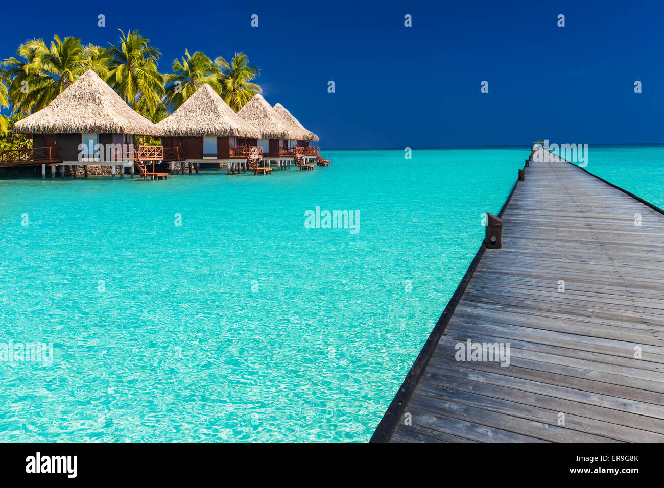 Long wodden jetty extended into azure water of lagoon with villas over the water Stock Photo