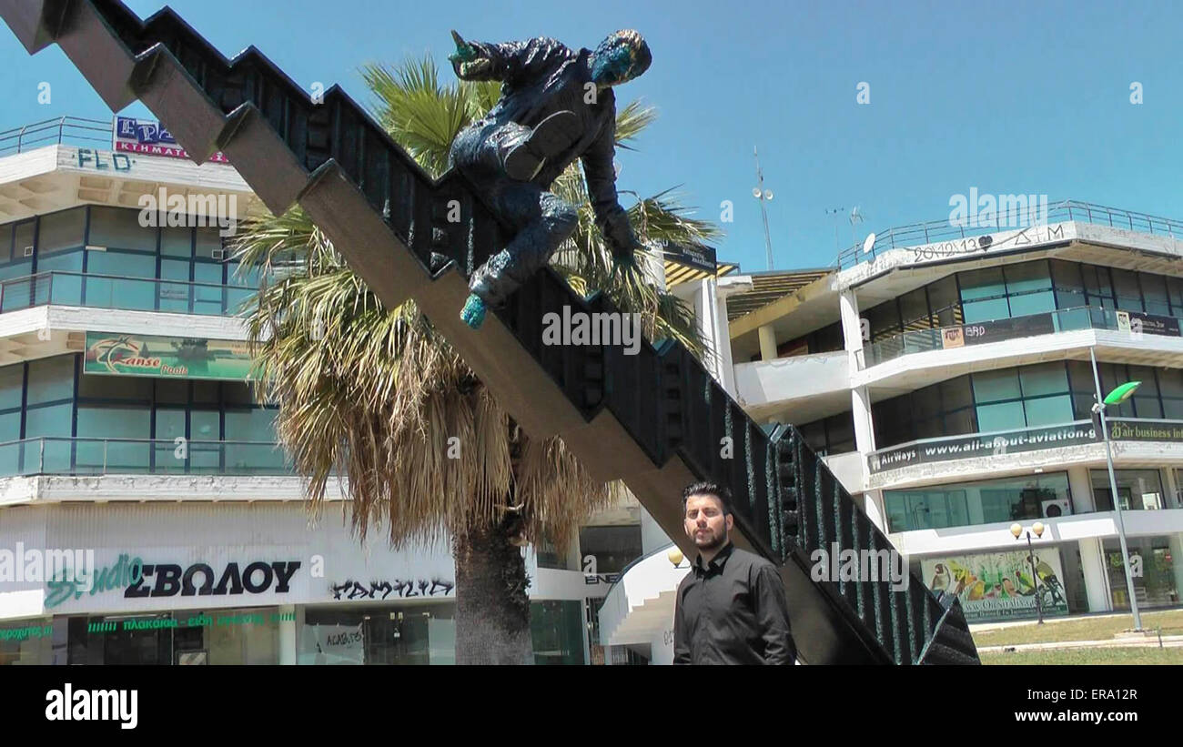 Athens. 14th May, 2015. Photo taken on May 14, 2015 shows 22-year-old Greek self-taught artist Tasos Nyfadopoulos posing for photos with his sculpture 'Crisis' in Athens, capital of Greece. Situated next to Vouliagmenis Avenue, a few kilometers from the center of Athens, the impressive sculpture, about 7 meters long and 4 meters high, is a giant stock exchange index that crushed on the ground and broke into pieces. On the top of it, a man is about to fall as the index collapses. © Athanasia Batziou/Xinhua/Alamy Live News Stock Photo