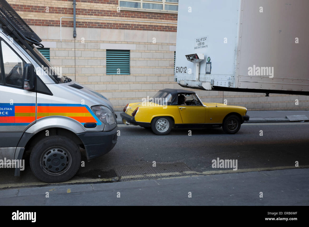 MGB Midget car drives past other parked vehicles, showing it's scale. It is so small it appears to be driving underneath a conta Stock Photo