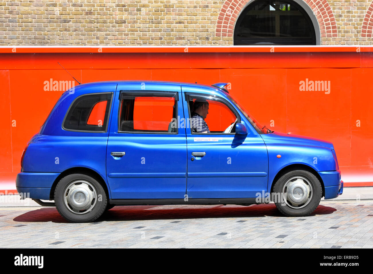 Unmarked London taxi cab driver reads newspaper queuing for customers at Kings Cross station Camden beside orange colour building site hoarding UK Stock Photo