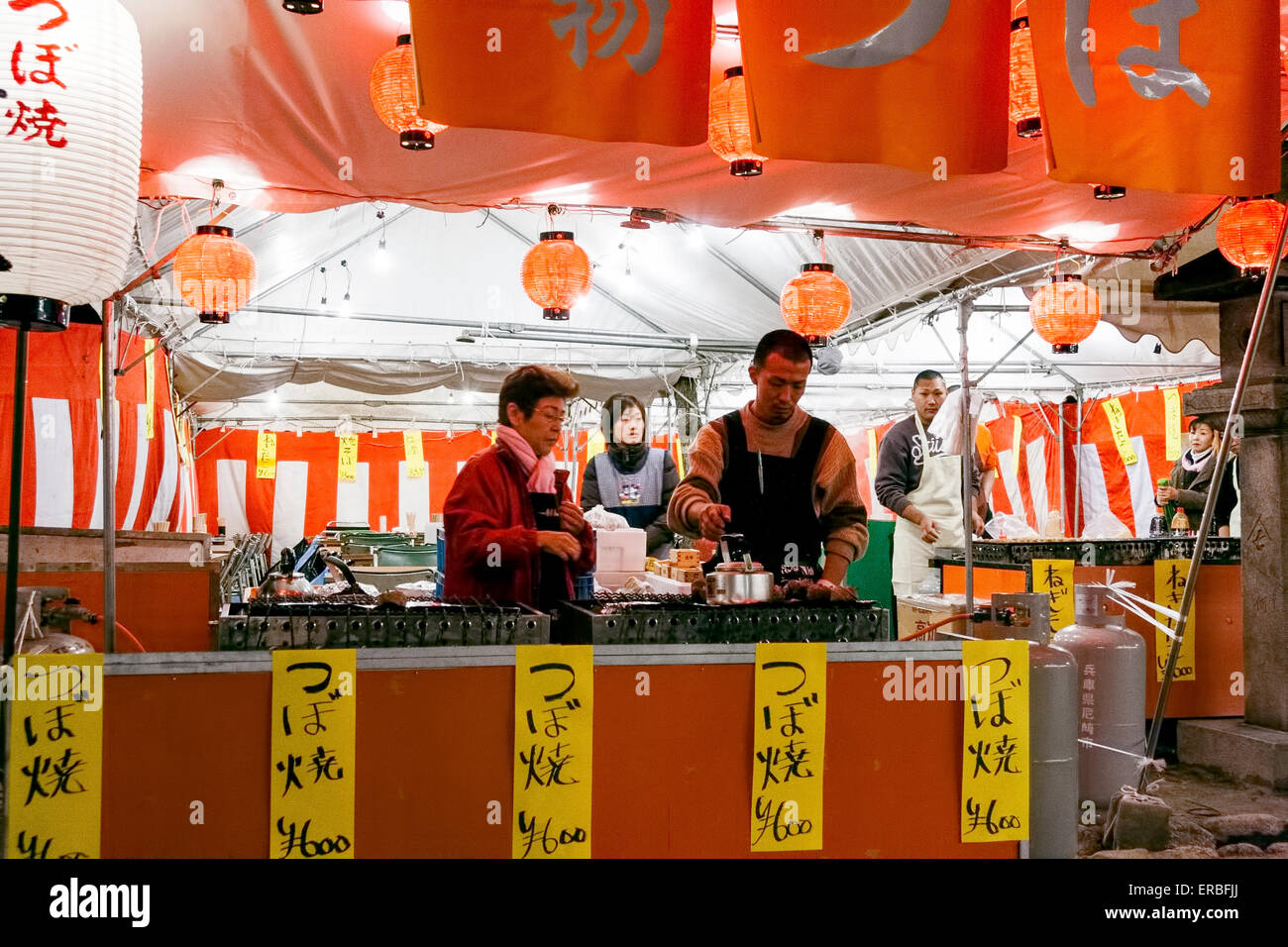 Japanese woman and man, facing, preparing tsuboyaki, turben shells, typical festival food, on a food stall, combined with others, at night. Stock Photo