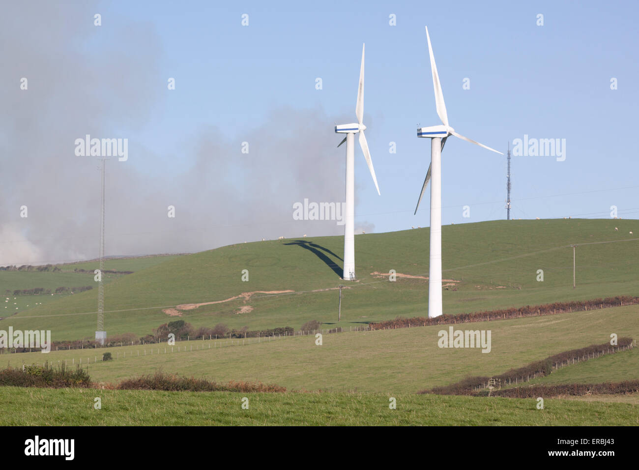 Two wind turbines in a field with smoke in the background Stock Photo