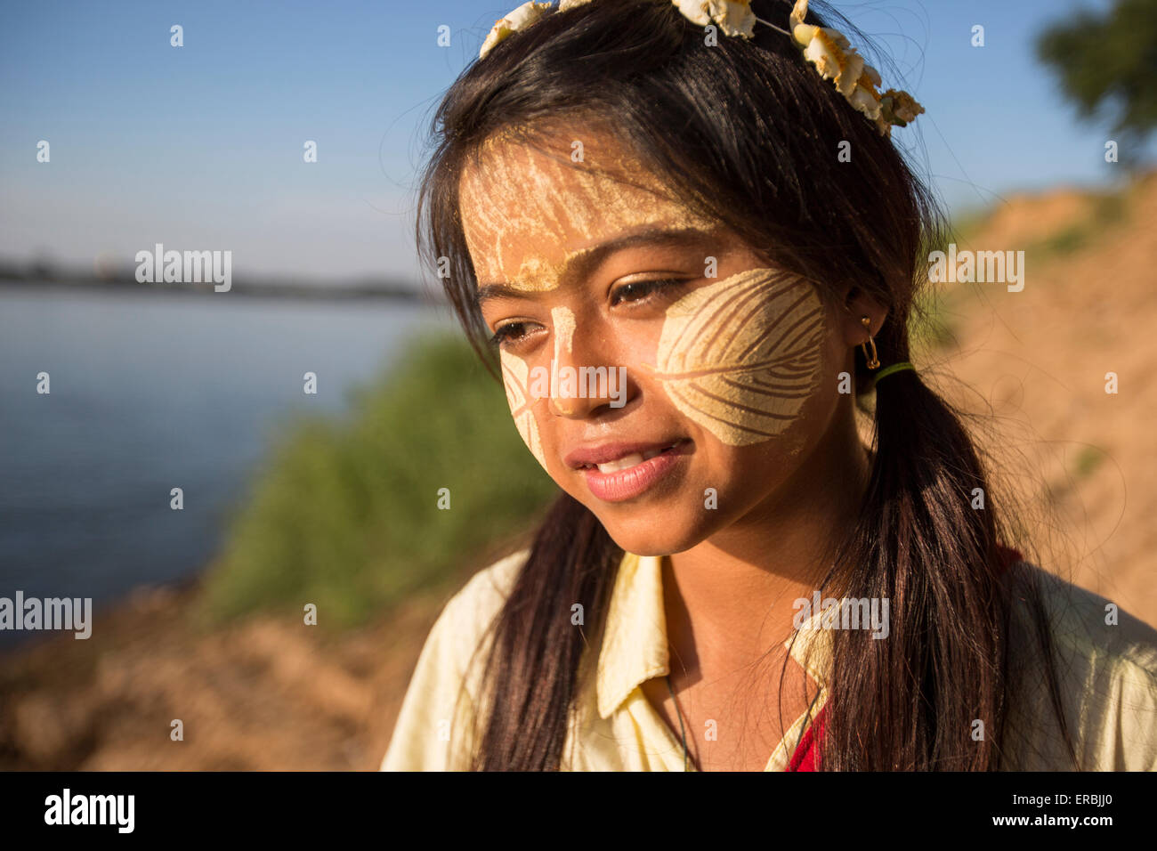Pretty young girl with gold leaf on face at water's edge of Ayeyarwady River, Myanmar Stock Photo