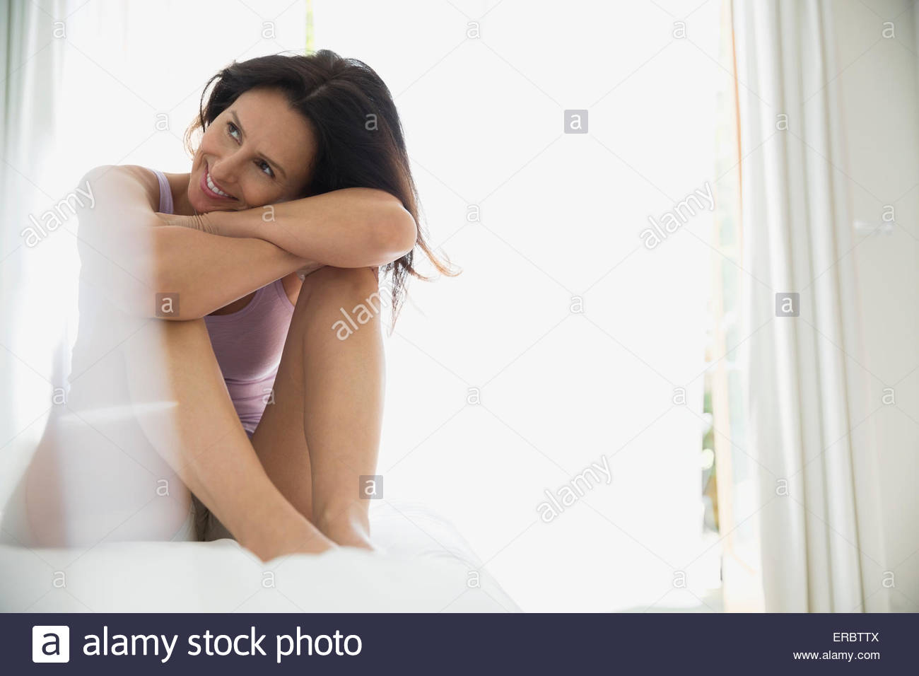 Smiling brunette woman hugging knees on bed Stock Photo