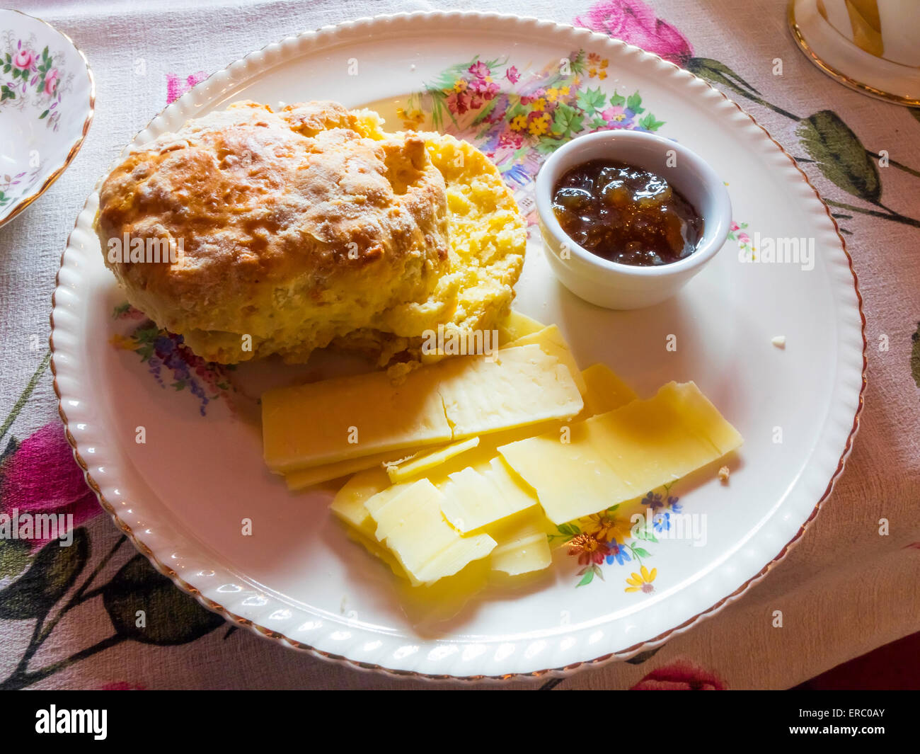 A serving of a Scone with Cheddar cheese slices and Chutney on a floral plate and tablecloth Stock Photo