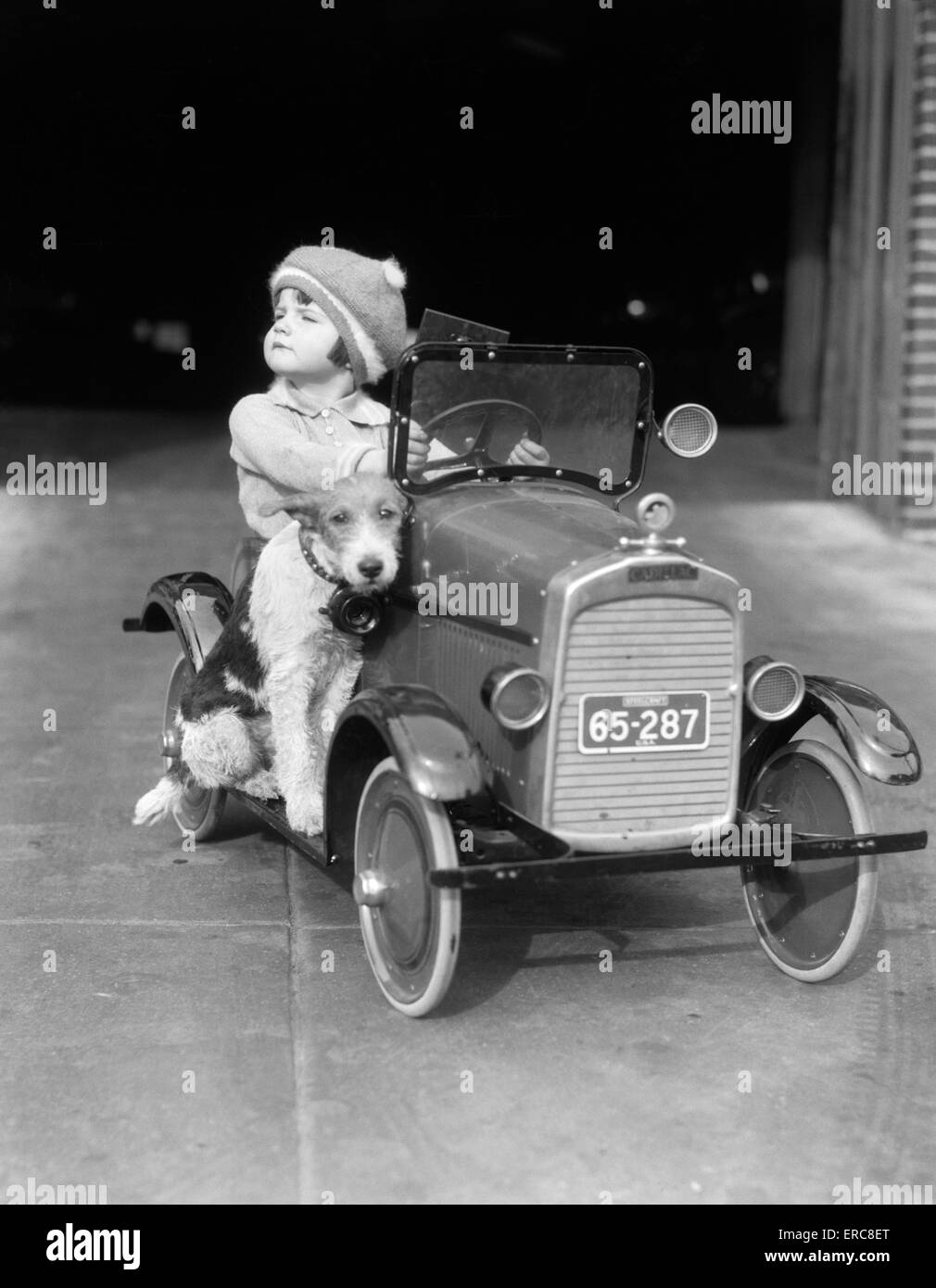 1920s GIRL IN TOY PEDAL CAR WITH DOG SITTING ON RUNNING BOARD GIRL LOOKING TO SIDE HEAD UP Stock Photo