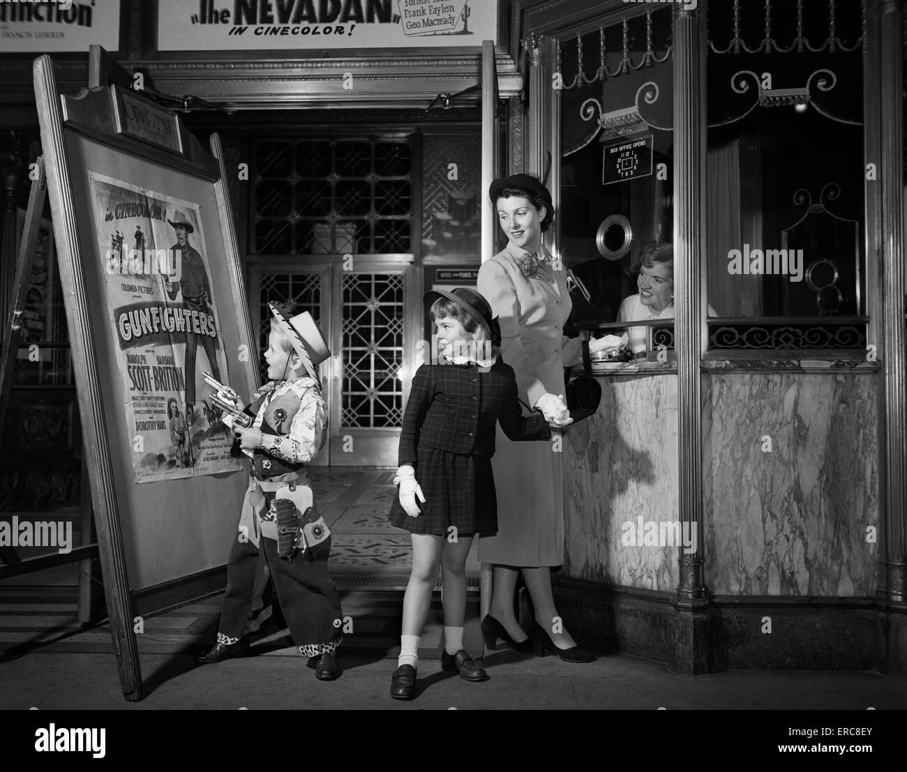 1950s MOTHER TAKING KIDS TO MOVIES BUYING TICKETS TO A WESTERN BOY WEARING COWBOY COSTUME Stock Photo
