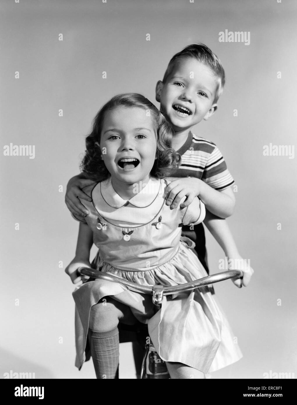 1950s GIRL WEARING A DRESS WITH  RIDING A TRICYCLE WHILE A BOY IS STANDING ON BACK HOLDING ON TO HER SHOULDERS Stock Photo