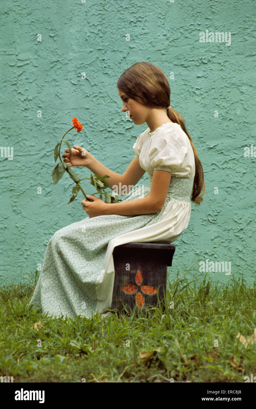 1960s PENSIVE YOUNG TEENAGE GIRL HOLDING LONG STEM FLOWER SITTING BENCH OUTDOORS PROFILE WEARING GRANNY DRESS Stock Photo
