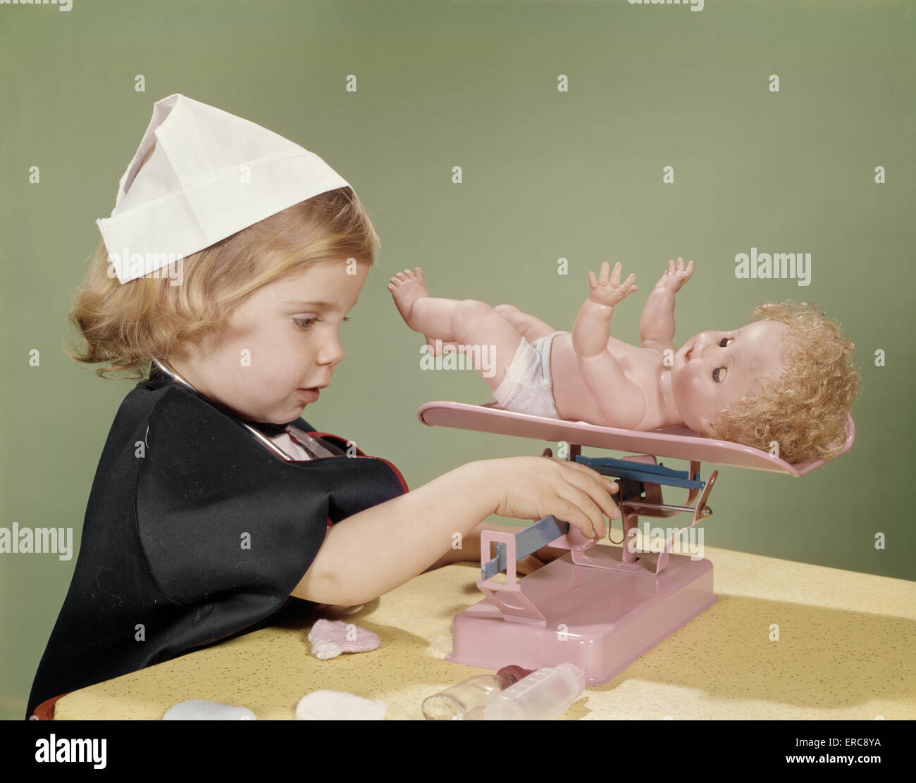 1960s LITTLE GIRL DRESSED IN NURSE'S COSTUME WEIGHING HER DOLL ON TOY BABY SCALE Stock Photo