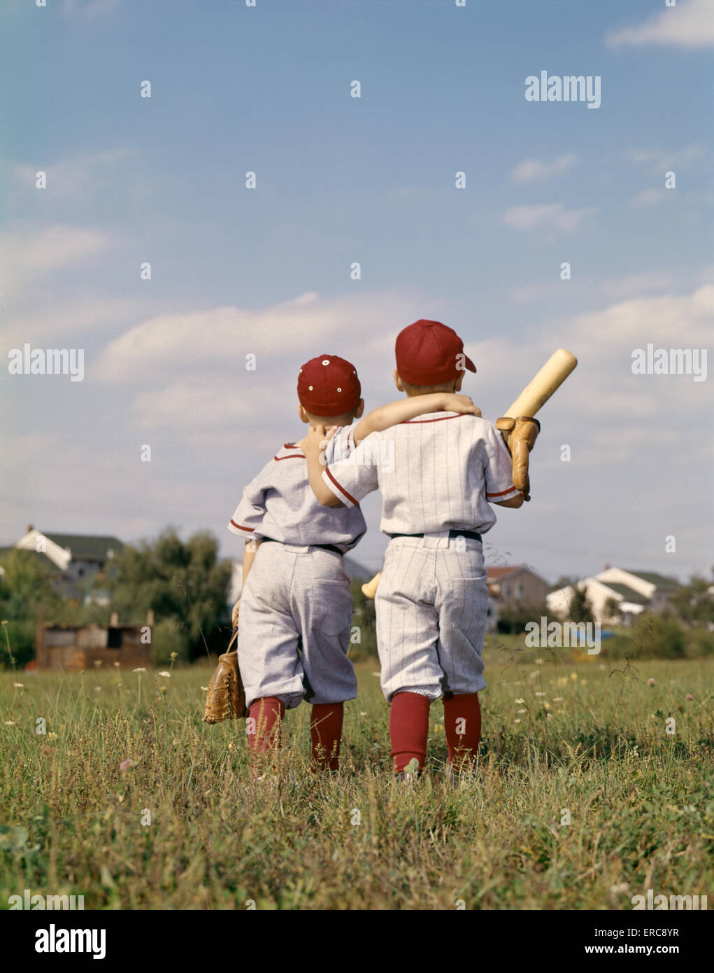 1960s TWO BOYS BROTHERS TEAMMATES WEARING LITTLE LEAGUE BASEBALL UNIFORMS WALKING ARM IN ARM AWAY FROM CAMERA Stock Photo