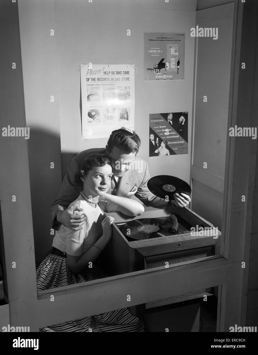 1950s COUPLE SITTING IN STORE RECORD LISTENING BOOTH MAN WITH ARM AROUND WOMAN HOLDING 78 RPM Stock Photo