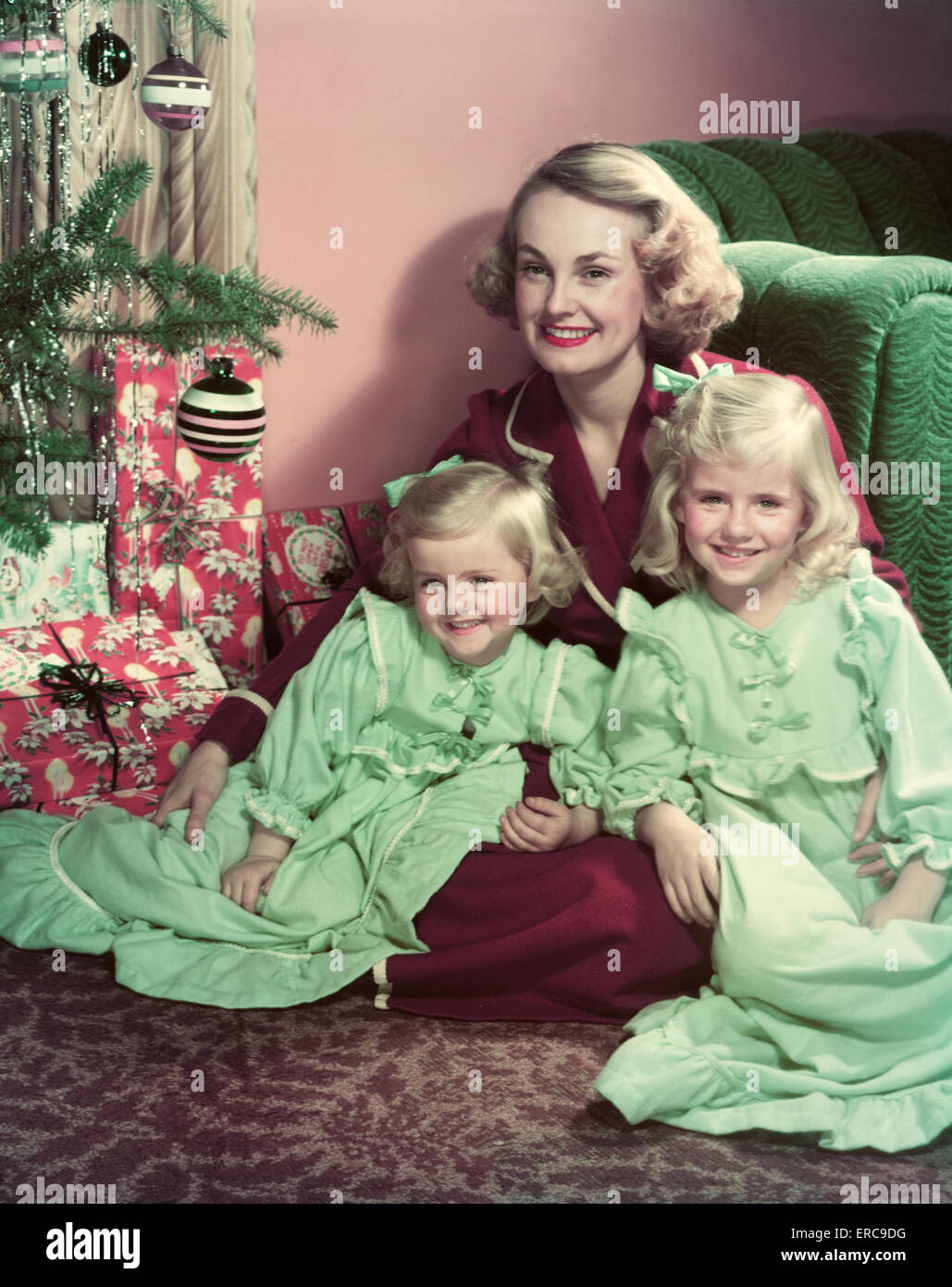 1950s SMILING MOTHER AND TWO DAUGHTERS IN NIGHTGOWNS POSING SITTING ON FLOOR BY CHRISTMAS TREE LOOKING AT CAMERA Stock Photo