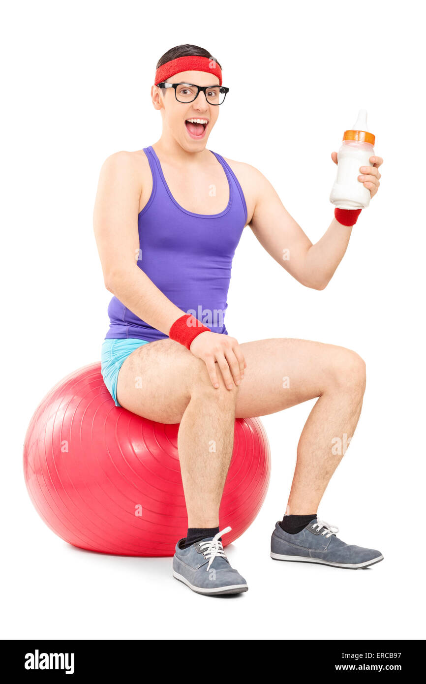 Vertical shot of a nerdy guy in sportswear sitting on a fitness ball and holding a huge baby bottle full of milk Stock Photo