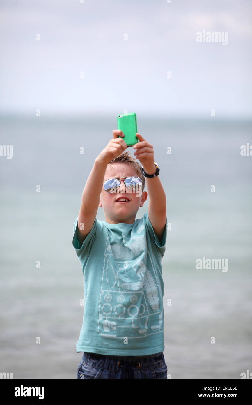 A boy wearing sunglasses on a beach taking a selfie photograph with his iPod to post onto instagram Stock Photo