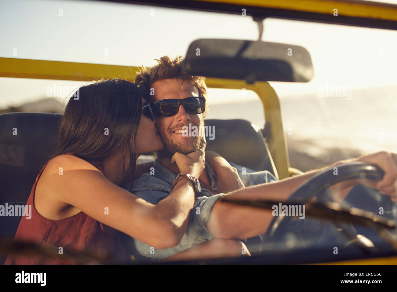 Loving young couple on road trip. Woman kissing her boyfriend's cheeks. Young man driving a car. Romantic young couple enjoying Stock Photo
