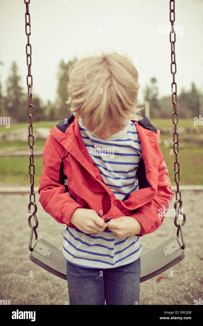 A young boy zips up his jacket before using the playground swing Stock
