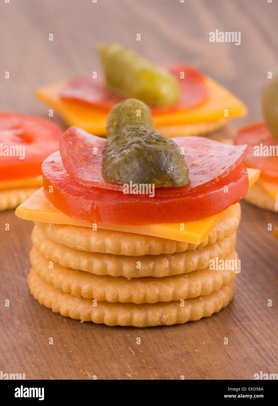 Stack of crackers, top one loaded with cheese, tomato, pepperoni and pickle; on dark wood table top Stock Photo