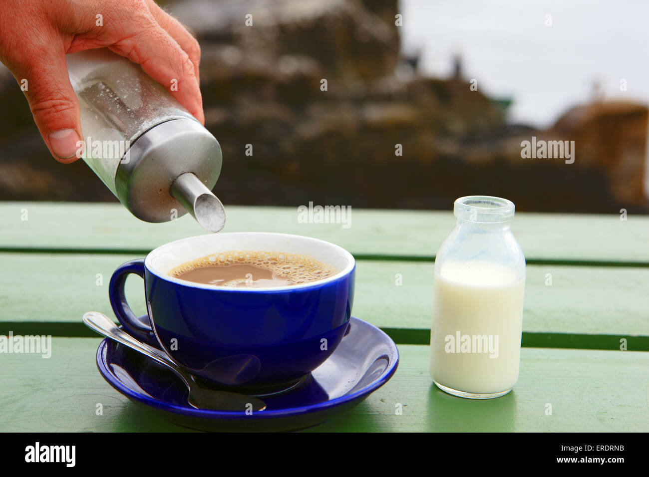 Cup of coffee on a wooden bench outside, having sugar poured into it and a small bottle of milk beside it Stock Photo