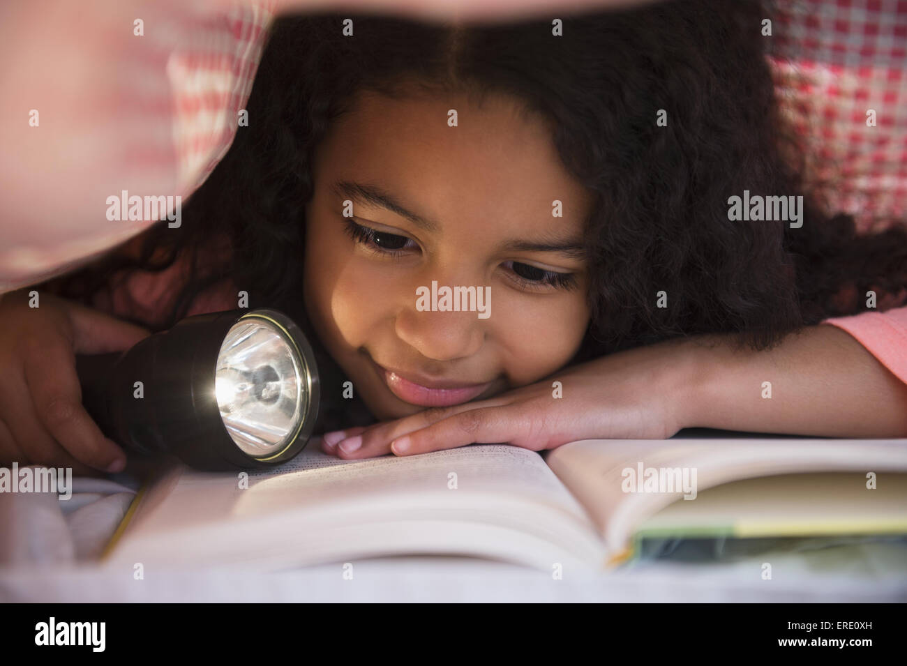 Mixed race girl reading book with flashlight under blanket Stock Photo