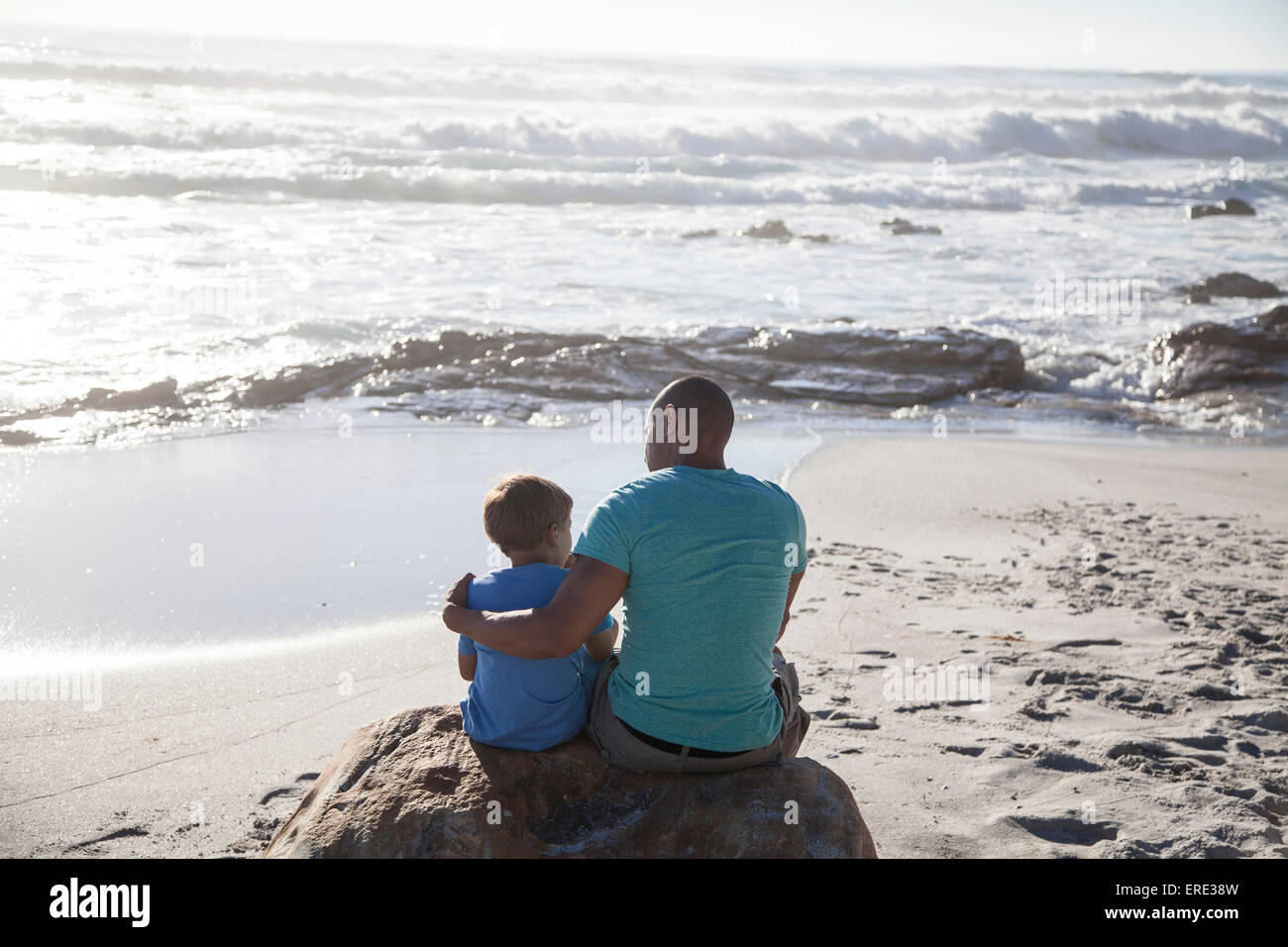 Mixed race father and son sitting on beach Stock Photo