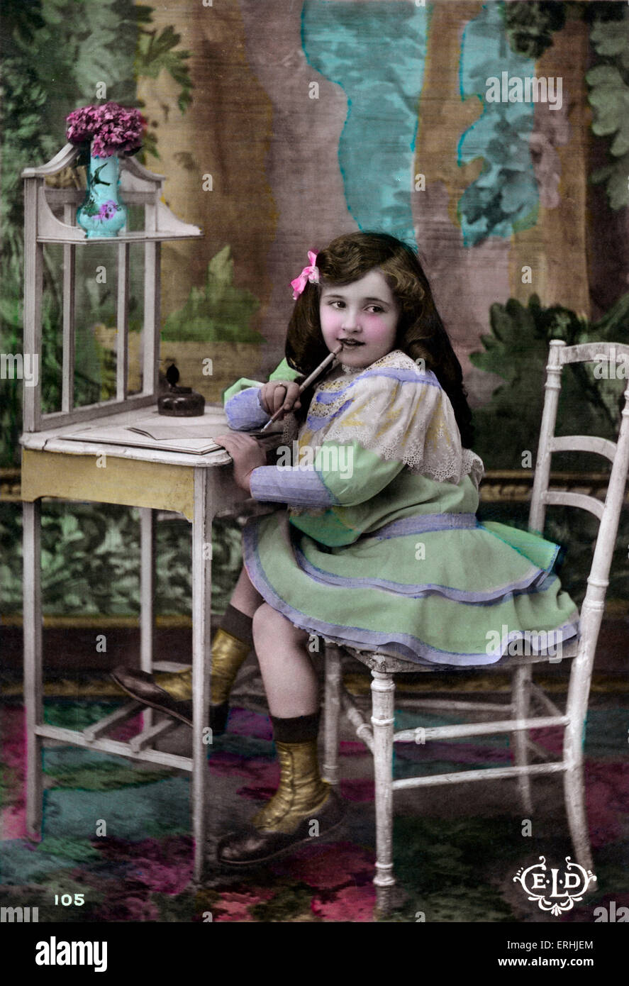 Girl writing New Year's letter. Hand coloured photograph. French post card. Writing desk. Dress. Boots. Pen and ink. Bow in Stock Photo