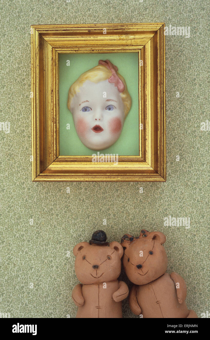 Two teddy bears standing against wallpapered wall beneath gold picture frame containing face of porcelain doll Stock Photo