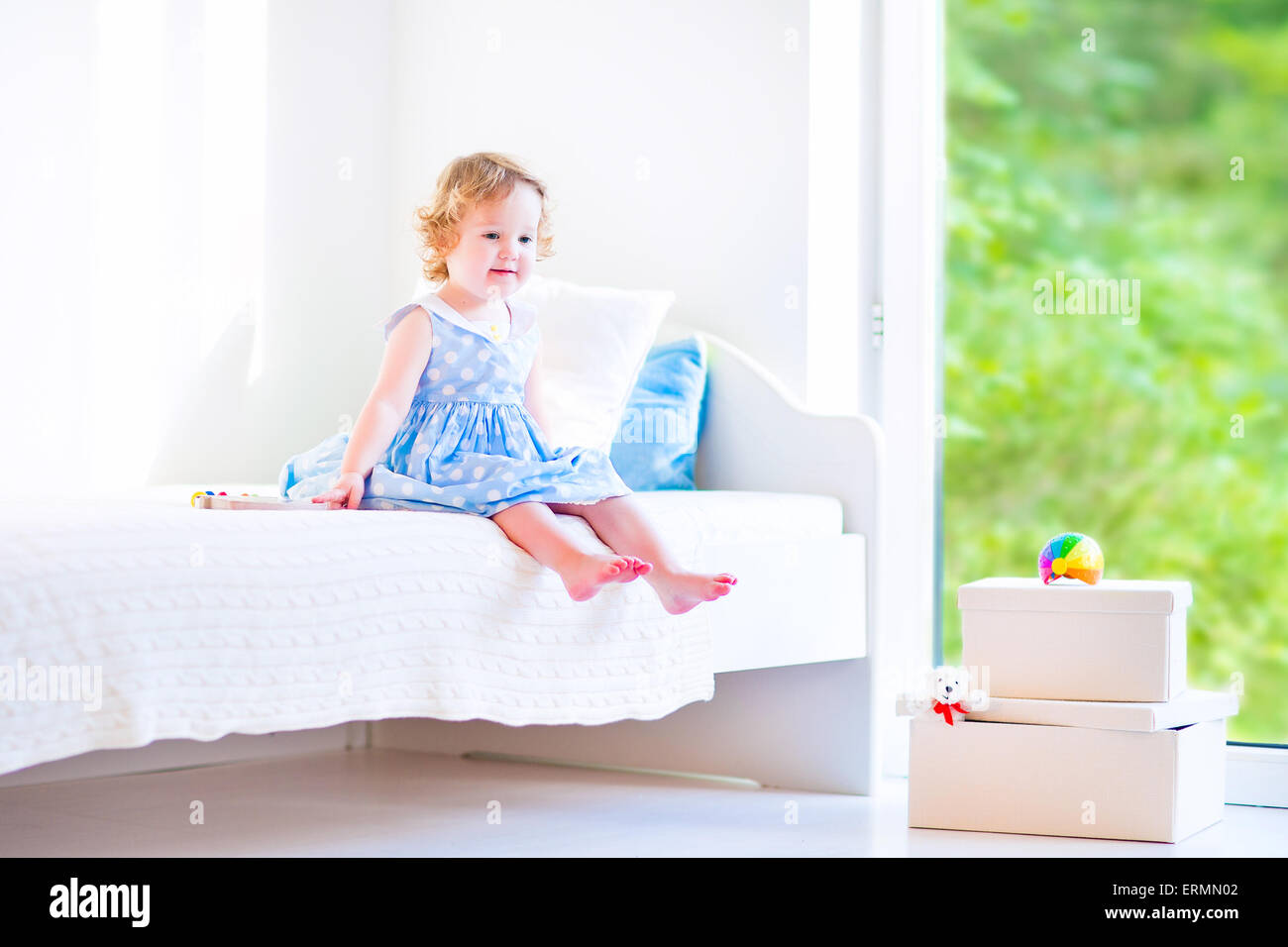 Cute toddler, adorable curly little girl in a blue dress, sitting on a bed reading a book playing in a sunny white bedroom Stock Photo